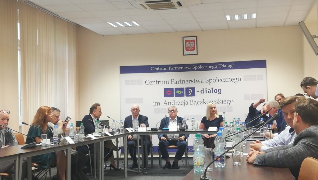 Przedstawiciele instytucji i organizacji publicznych siedzą przy stołach ustawionych w koło. Na ścianie baner Centrum Partnerstwa Społecznego – dialog, w którym odbyło się spotkanie.