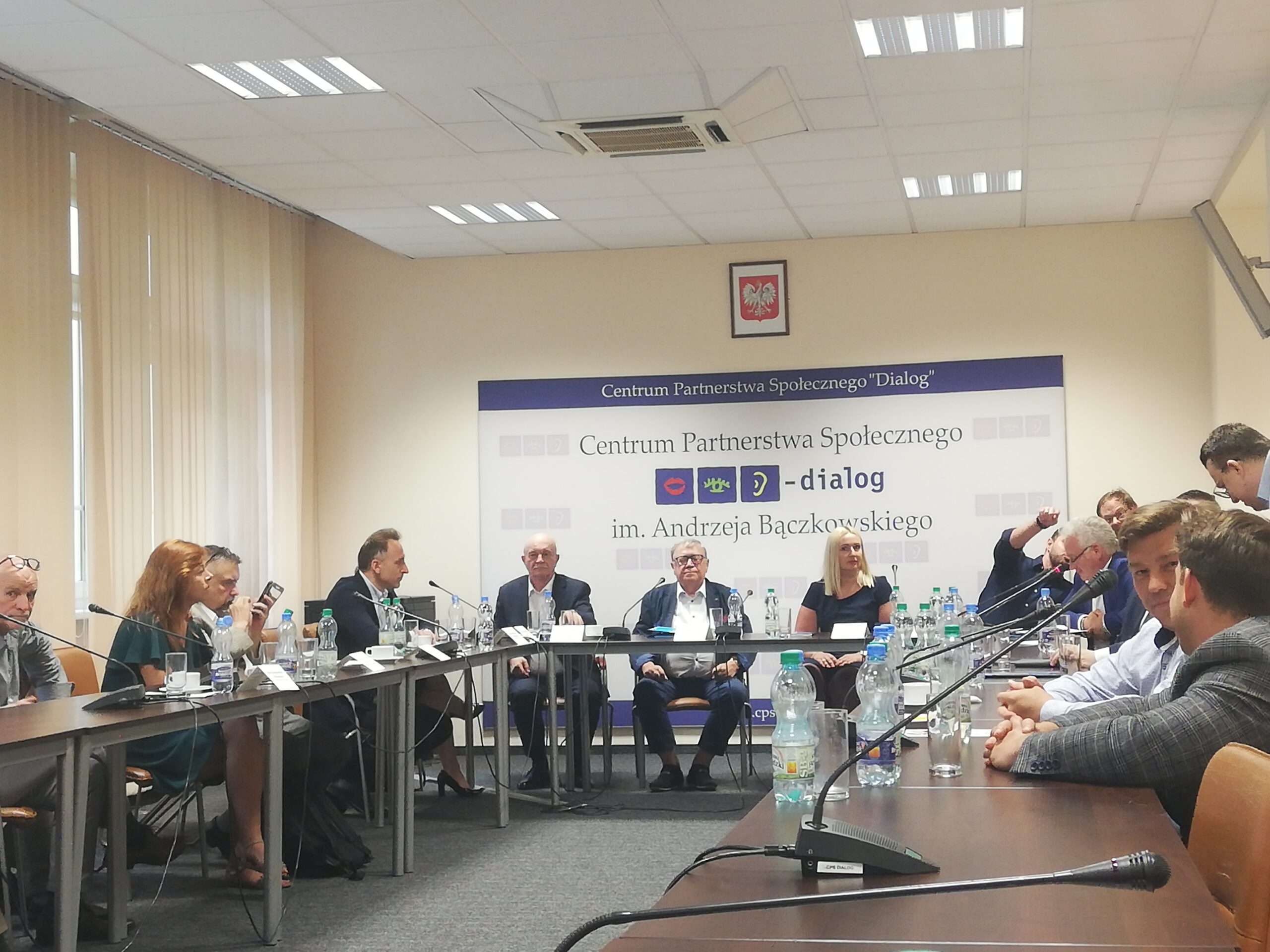 Przedstawiciele instytucji i organizacji publicznych siedzą przy stołach ustawionych w koło. Na ścianie baner Centrum Partnerstwa Społecznego – dialog, w którym odbyło się spotkanie.