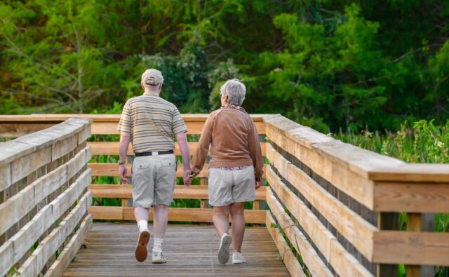 Dwoje seniorów, kobiet i mężczyzna idą po drewnianym mostku, wkoło drzewa