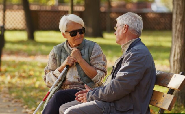 Seniorka trzymająca białą laskę siedzi na ławce i rozmawia ze starszym mężczyzną