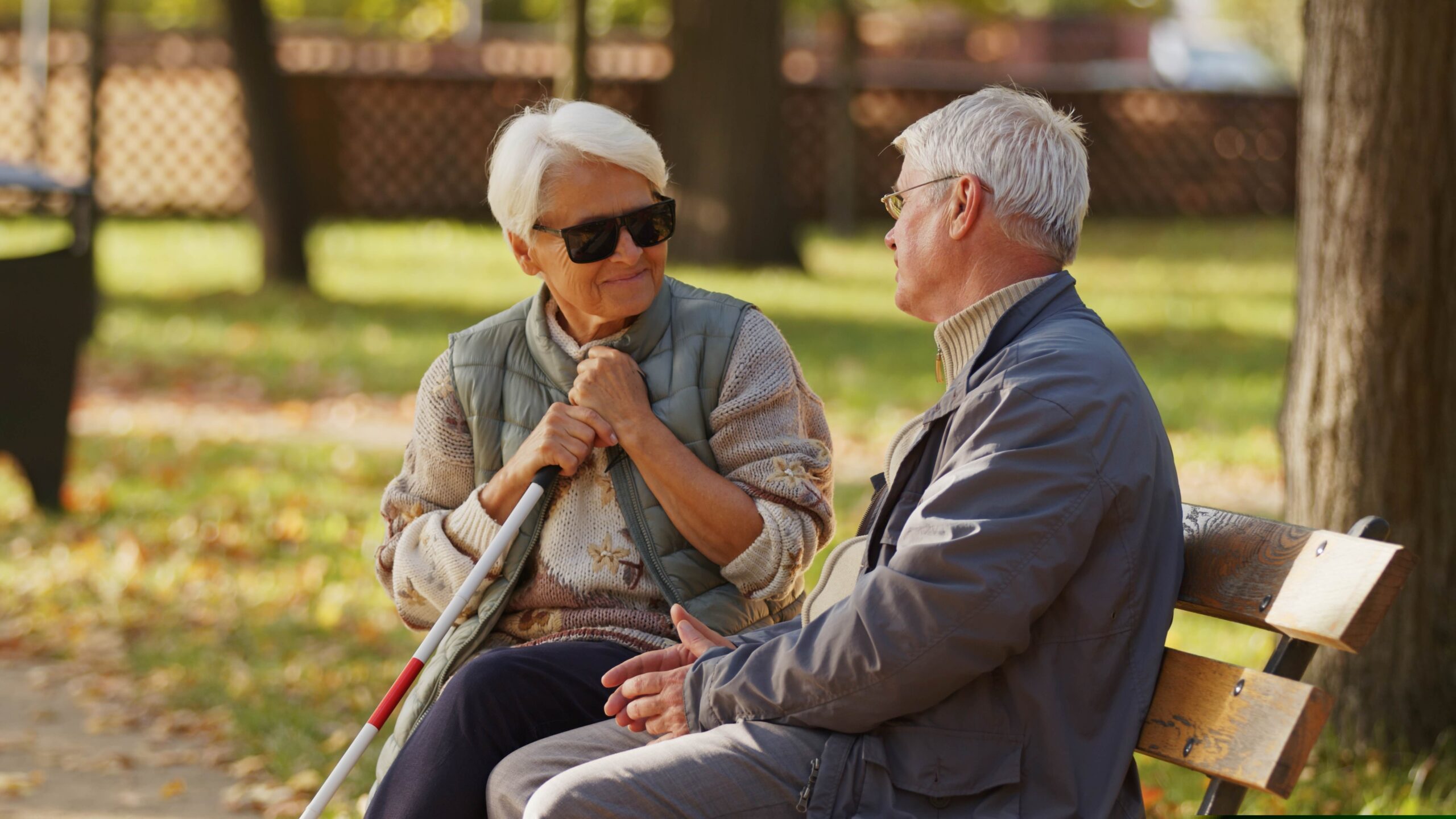 Seniorka trzymająca białą laskę siedzi na ławce i rozmawia ze starszym mężczyzną