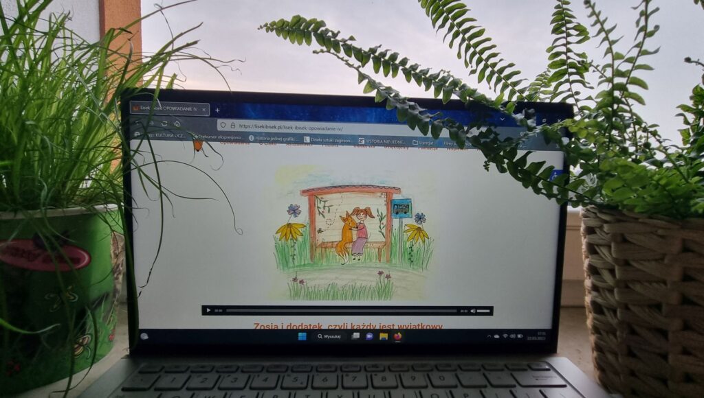 laptop stoi na parapecie, za nim jest okno, po jego lewej i prawej stronie stoi kwiatek, na ekranie komputera widać stronę z opowiadaniami o Lisku Ibisku