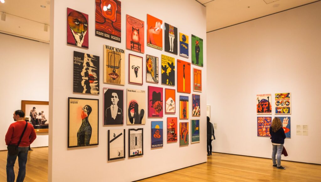 Wnętrze galerii sztuki. Na ścianach kolorowe grafiki. Oglądają je pojedynczy goście galerii.