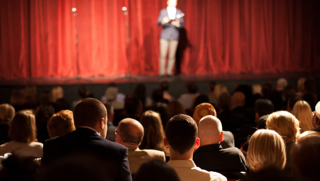 Teatr, Pełna widownia, przed nimi scena. Przed nieodsłoniętą jeszcze kurtyną sceniczną stoi mówca.