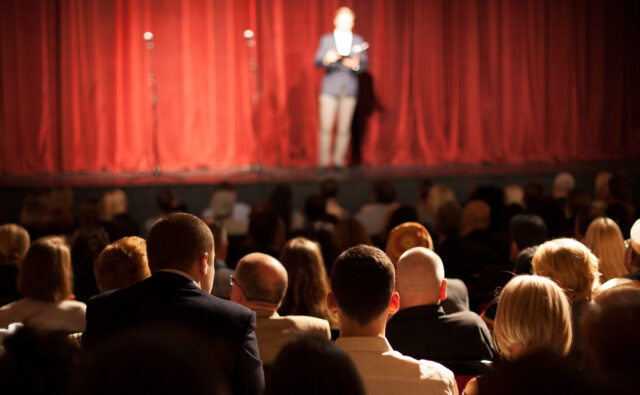 Teatr, Pełna widownia, przed nimi scena. Przed nieodsłoniętą jeszcze kurtyną sceniczną stoi mówca.