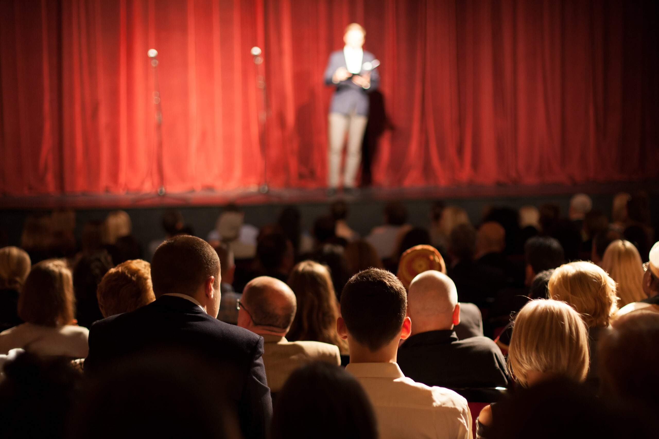 Teatr, Pełna widownia, przed nimi scena. Przed nieodsłoniętą jeszcze kurtyną sceniczną stoi mówca.