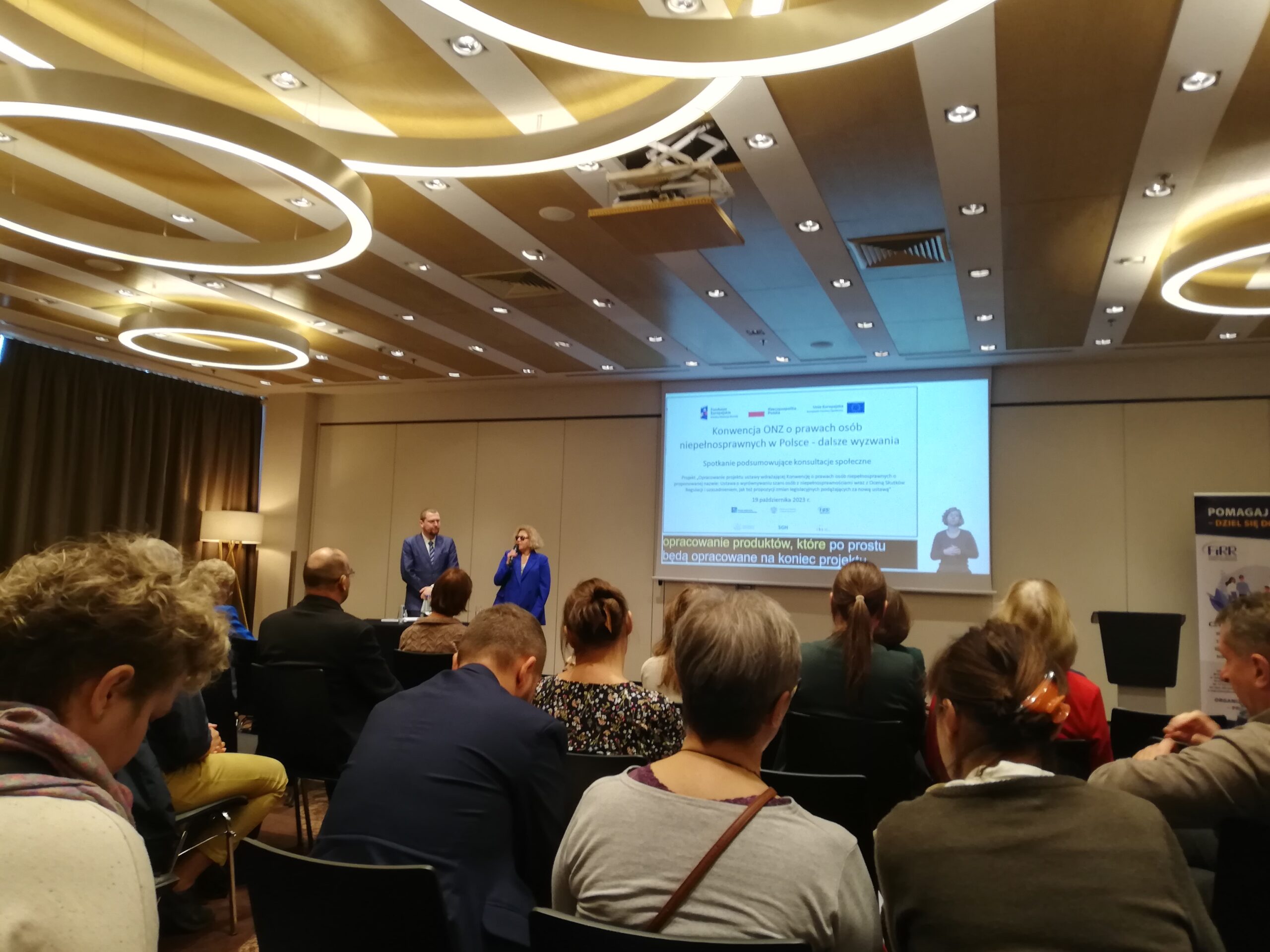 Na pierwszym planie siedzą uczestnicy konferencji. Na drugim planie dwóch prowadzących mówi do mikrofonu.