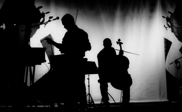 Na scenie widać czarne figury kontrabasisty i muzyka trzymającego nuty, w tle światło reflektorów.