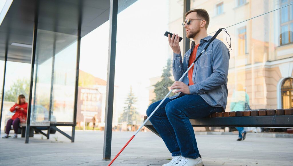 Niewidomy mężczyzna siedzi na ławce mówi przez telefon, w ręce trzyma białą laskę.