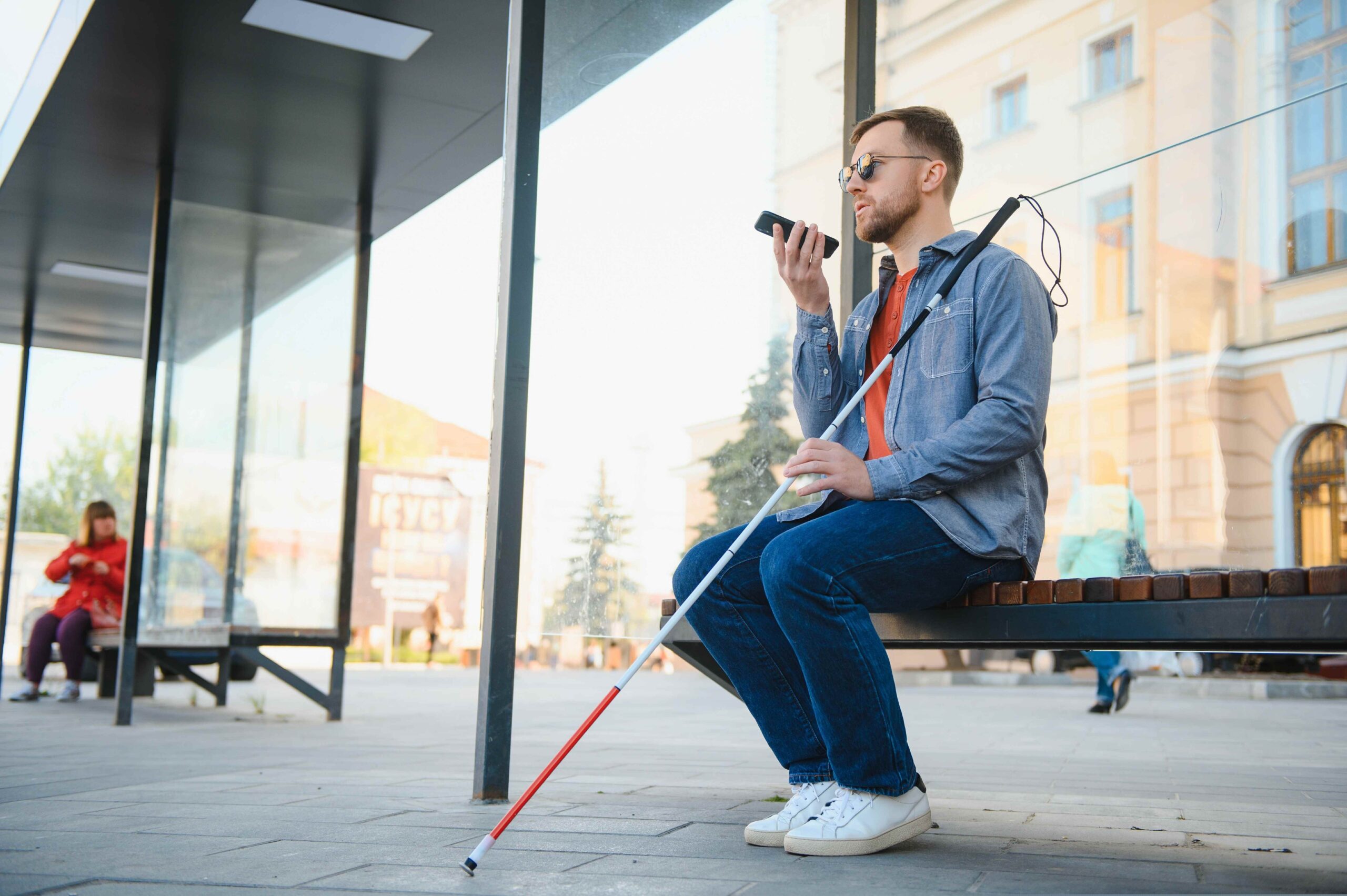 Niewidomy mężczyzna siedzi na ławce mówi przez telefon, w ręce trzyma białą laskę.