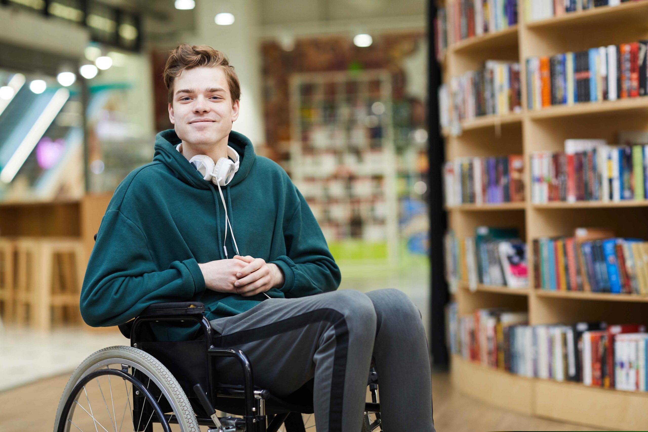 Młody chłopak na wózku inwalidzkim uśmiecha się, w tle biblioteka.