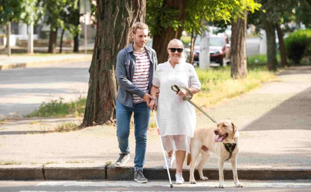 Młody chłopak przeprowadza niewidomą kobietę przez ulicę.