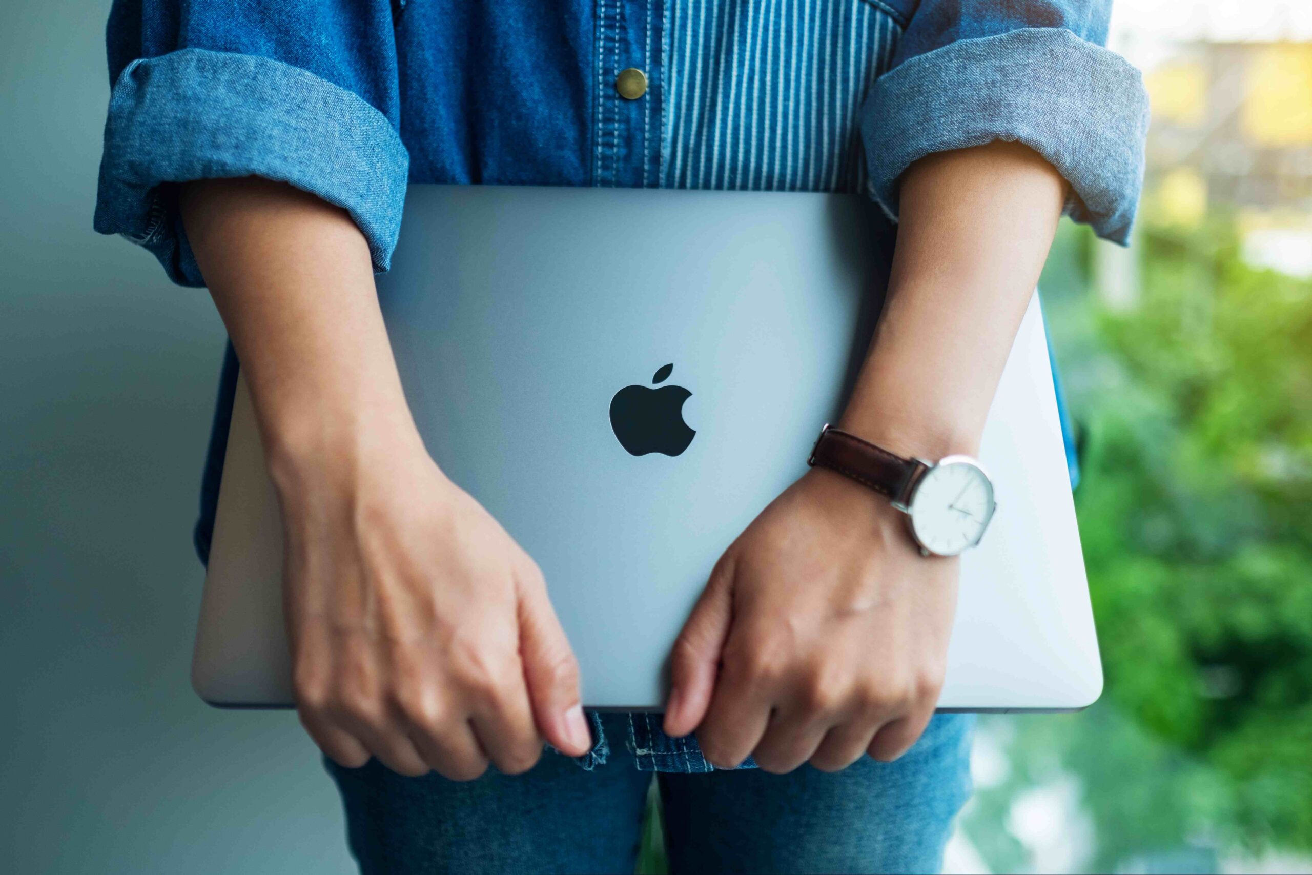 Zamknięty laptop trzymany przed osobą. Widoczne ręce i klapa laptopa z symbolem jabłka firmy Apple.