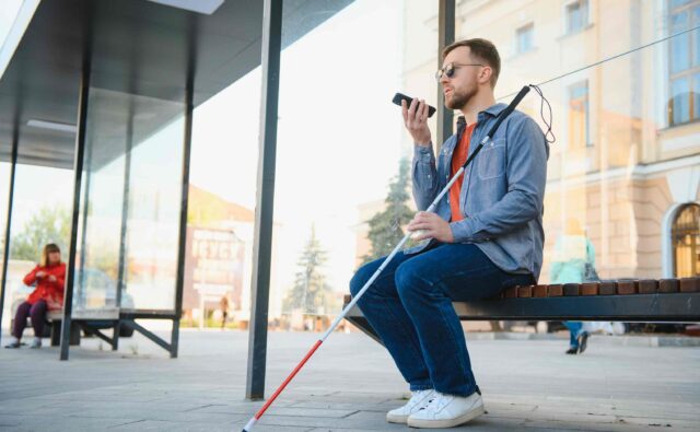 Młody mężczyzna białą laską siedzi na ławce na przystanku. w ręku trzyma smartfon.