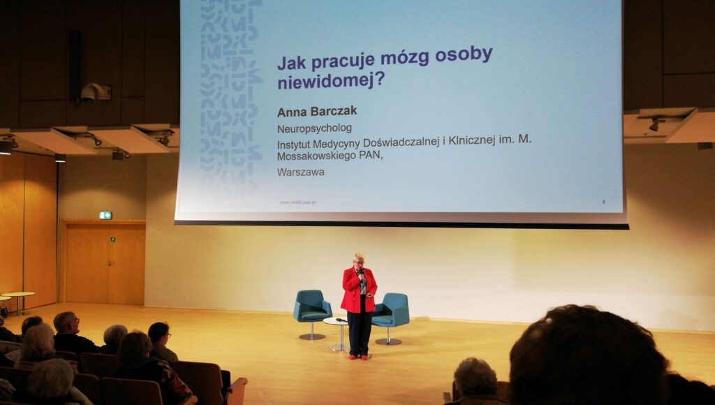 Sala wykładowa. Rzędy siedzeń w ustawieniu kinowym. Przed widzami prelegentka. Blondynka z krótkimi włosami w średnim wieku. Okulary z czarnymi oprawkami, czerwone marynarka i buty, czarne spodnie, w dłoni mikrofon. Nad nią wielki ekran z tytułowym slajdem prezentacji. Napis „Jak pracuje mózg osoby niewidomej?” pod spodem informacje o autorce: Anna Barczak Neuropsycholog Instytut Medycyny Doświadczalnej i Klinicznej im. M. Mossakowskiego PAN Warszawa