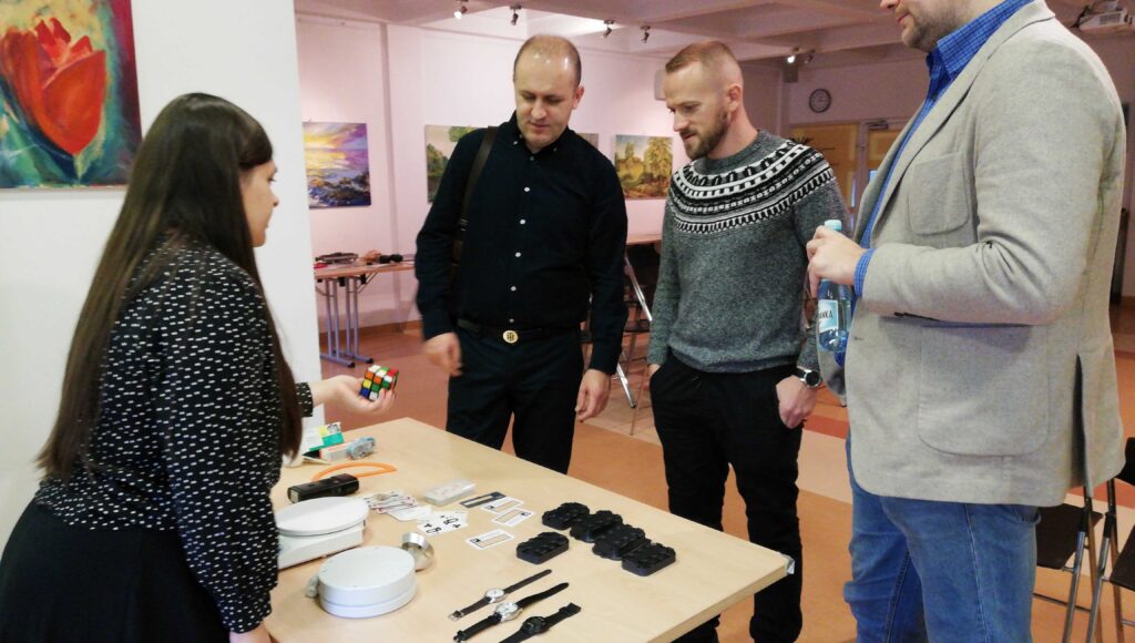 Trzech mężczyzn stoi przy stoliku, na którym leżą, m.in. zegarki brajlowskie, waga z funkcją głosową, ramka do podpisu i inne. Przed nimi kobieta trzyma w dłoni, skierowanej w ich stronę, dotykową kostkę Rubika.