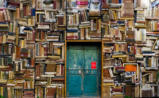 Biblioteka pełna książek, ściana z książkami od góry do dołu a pośrodku drzwi wejściowe.