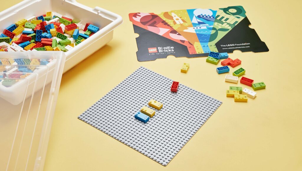 Klocki LEGO Braille Bricks ułożone na specjalnej planszy. Obok inne klocki tego typu oraz opakowanie tych klocków.