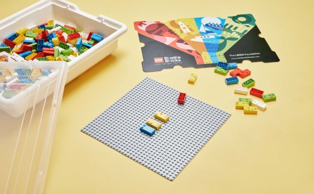 Klocki LEGO Braille Bricks ułożone na specjalnej planszy. Obok inne klocki tego typu oraz opakowanie tych klocków.