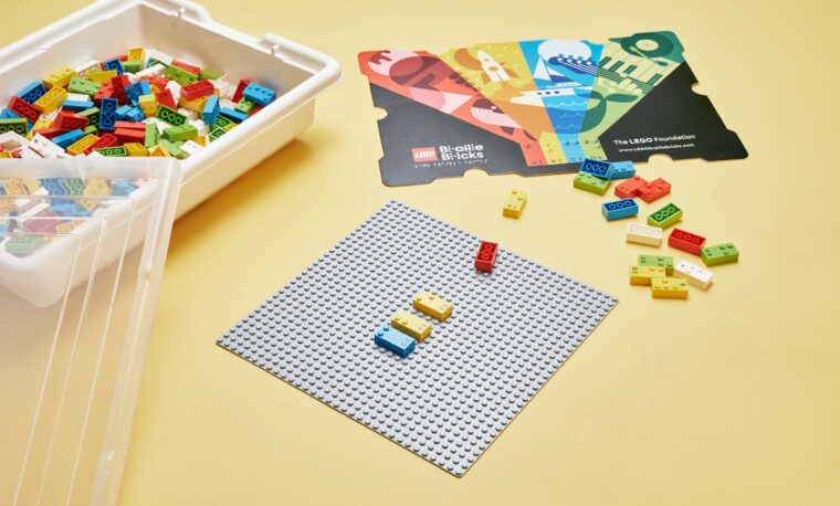 Klocki LEGO Braille Bricks ułożone na specjalnej planszy. Obok inne klocki tego typu oraz opakowanie tych klocków.