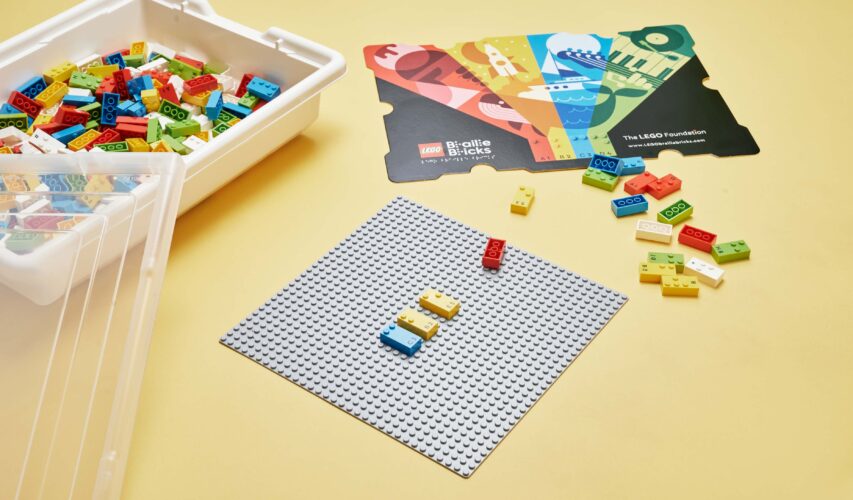 Klocki LEGO Braille Bricks ułożone na specjalnej planszy. Obok inne klocki tego typu oraz opakowanie tych klocków.