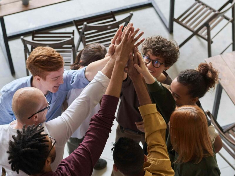 Zespół uśmiechniętych osób łączy prawe ręce wyciągnięte w górę w geście współpracy. Scena we wnętrzu, przy stołach z krzesłami.