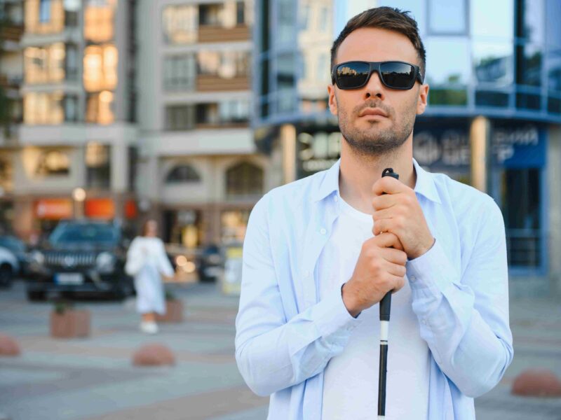 Niewidomy mężczyzna z białą laską stoi przed szklanym biurowcem, w którym znajdują się różne firmy.