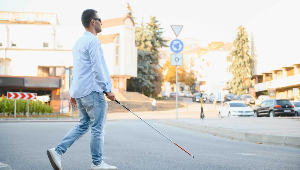 Młody niewidomy chłopak z białą laską przechodzi przez ulicę. W tle miejskie ulice.