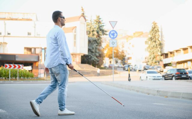 Młody niewidomy chłopak z białą laską przechodzi przez ulicę. W tle miejskie ulice.