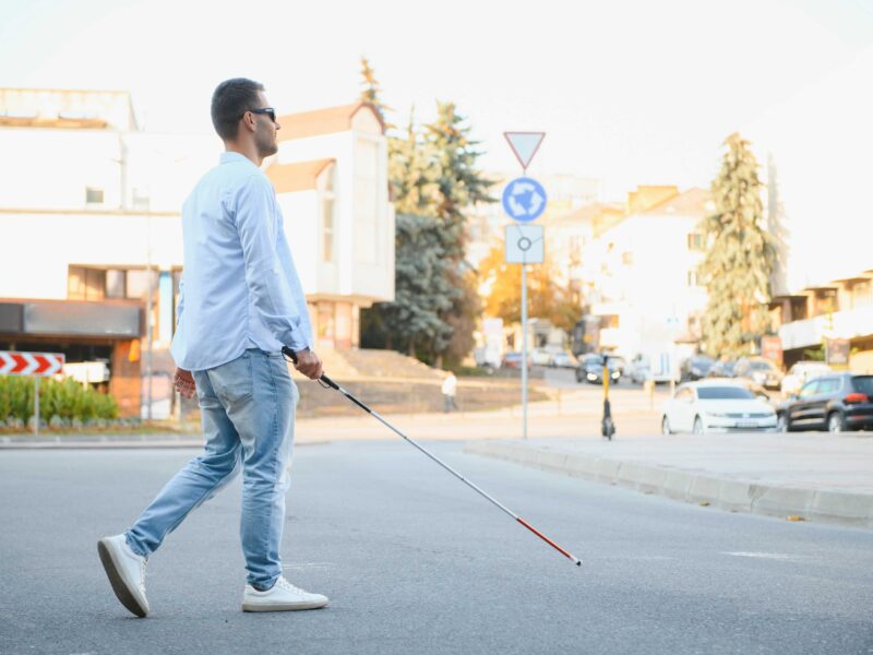 Młody niewidomy chłopak z białą laską przechodzi przez ulicę. W tle miejskie ulice.