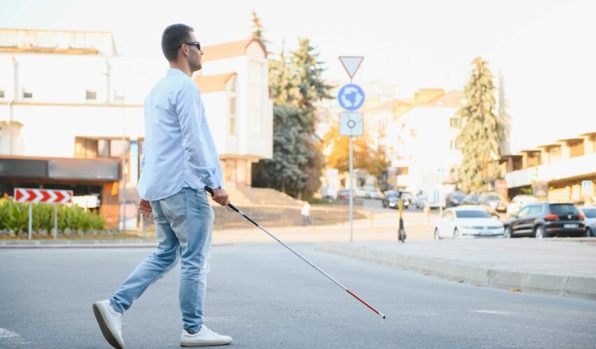 Młody niewidomy chłopak z białą laską przechodzi przez ulicę. W tle miejskie ulice.