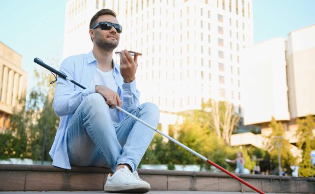 Młody niewidomy chłopak siedzi w centrum miasta i rozmawia przez smartfon.
