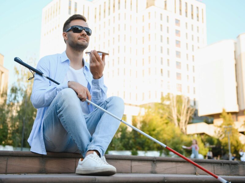 Młody niewidomy chłopak siedzi w centrum miasta i rozmawia przez smartfon.