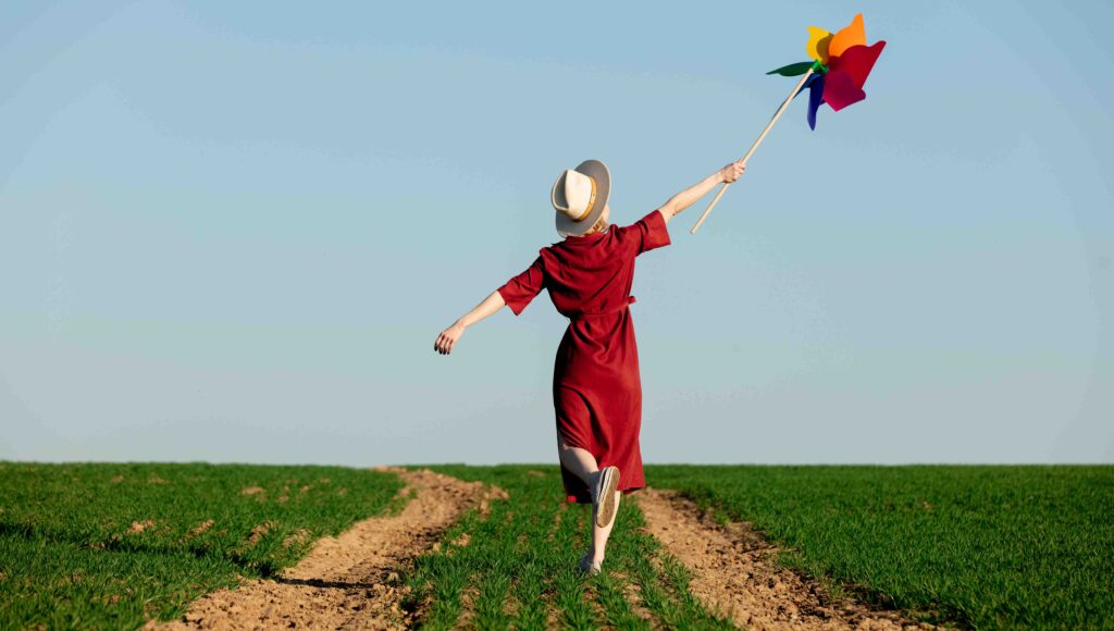 Kobieta idzie po polu w ręku trzyma papierowy wiatraczek. Jest odwrócona.