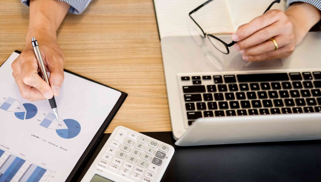 Osoba pracuje na laptopie obok kartka w wykresem i kalkulator.