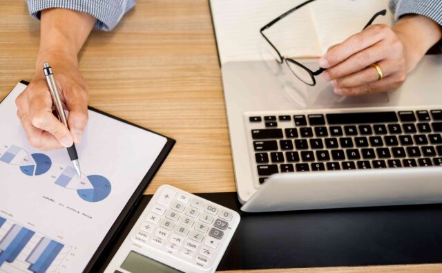 Osoba pracuje na laptopie obok kartka w wykresem i kalkulator.