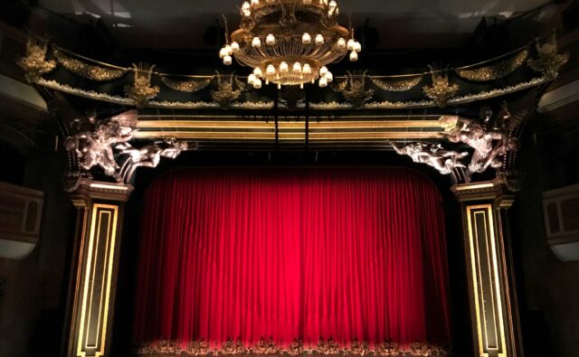 Czerwona kurtyna w teatrze, w tle złocenia i złoty ozdobny żyrandol.