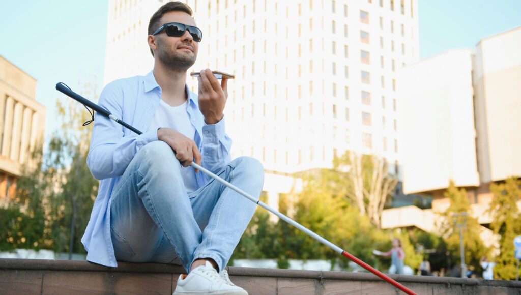 Młody niewidomy chłopak z białą laską siedzi n murku, w tle widać centrum miasta.