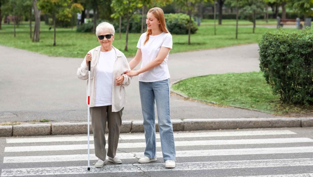 Młoda kobieta pomaga przejść przez przejście dla pieszych niewidomej kobiecie, seniorce, z białą laską.
