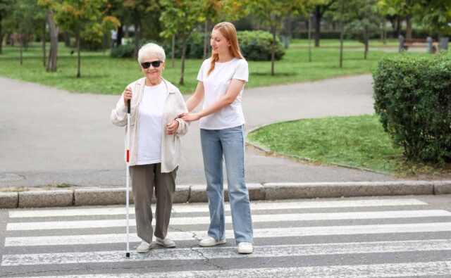 Młoda kobieta pomaga przejść przez przejście dla pieszych niewidomej kobiecie, seniorce, z białą laską.