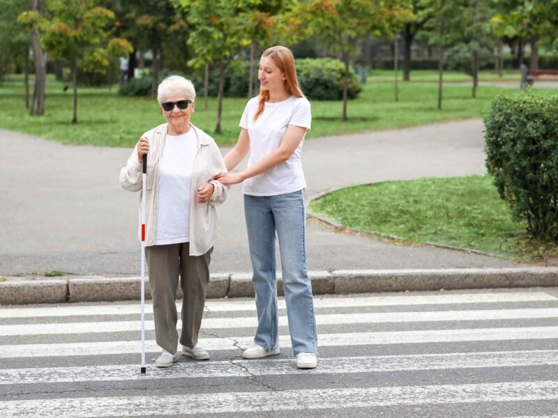Młoda kobieta pomaga przejść przez przejście dla pieszych niewidomej kobiecie, seniorce, z białą laską.