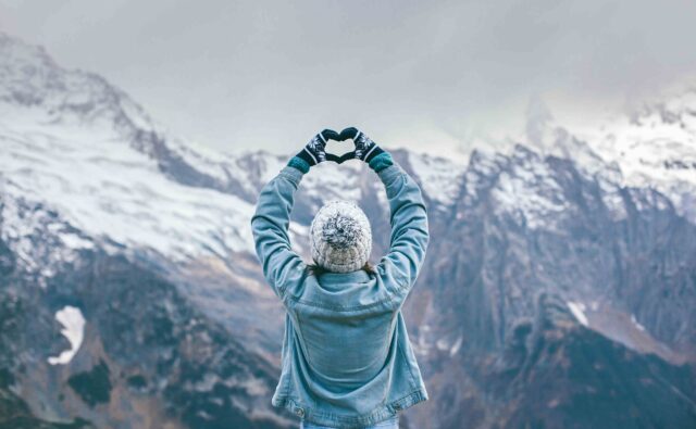 Dziewczyna robi znak serca dłońmi. W tle ośnieżone szczyty gór.