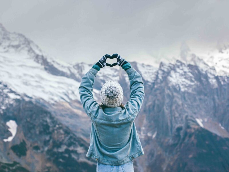 Dziewczyna robi znak serca dłońmi. W tle ośnieżone szczyty gór.