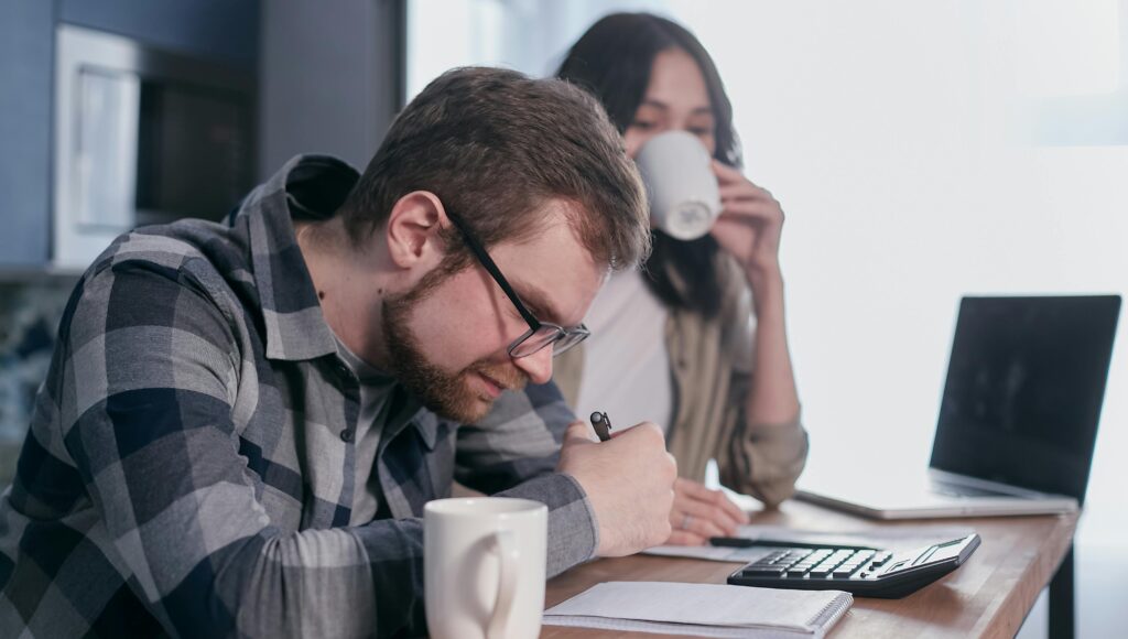 Mężczyzna w okularach liczy na kalkulatorze przy stole. Na stole kubek, laptop i dokumenty. Obok kobieta pije kawę.