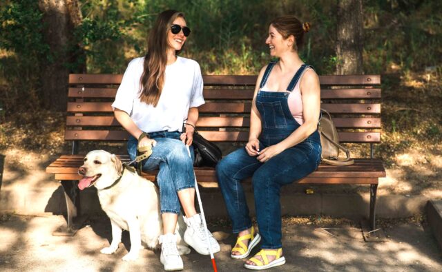 Młoda dziewczyna z białą laską i psem przewodnikiem siedzi na ławce i rozmawia z drugą młodą dziewczyną. Uśmiechają się obie.