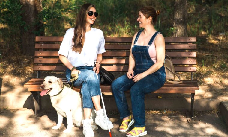 Młoda dziewczyna z białą laską i psem przewodnikiem siedzi na ławce i rozmawia z drugą młodą dziewczyną. Uśmiechają się obie.