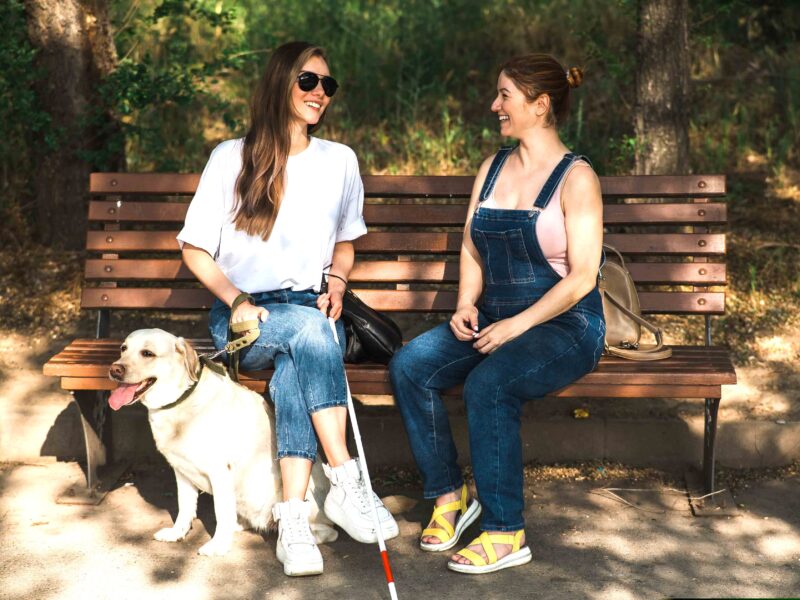 Młoda dziewczyna z białą laską i psem przewodnikiem siedzi na ławce i rozmawia z drugą młodą dziewczyną. Uśmiechają się obie.