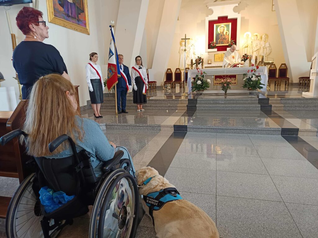 Osoba na wózku inwalidzkim z psem przewodnikiem uczestniczy w mszy św. W oddali ołtarz.