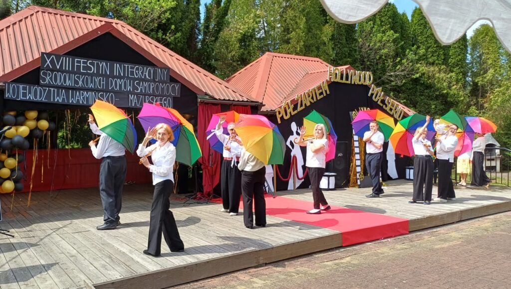 Uczestnicy festynu z kolorowymi parasolkami wykonują układ taneczny.