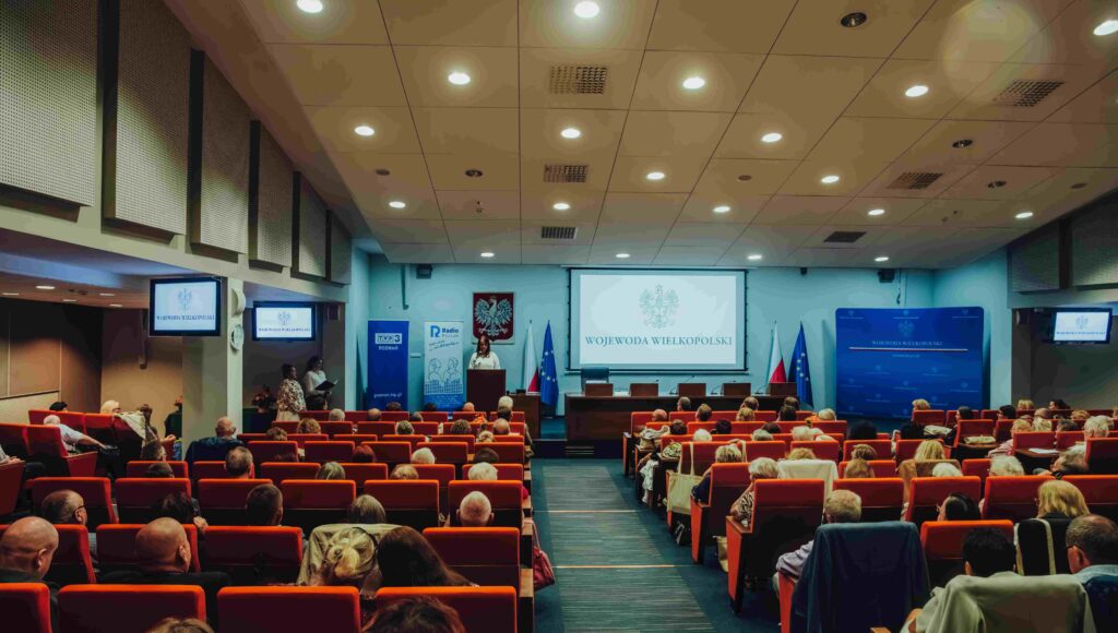 W oddali scena, a tam osoba przemawiająca do uczestników Jubileuszowej Konferencji Okręgu Wielkopolskiego PZN.